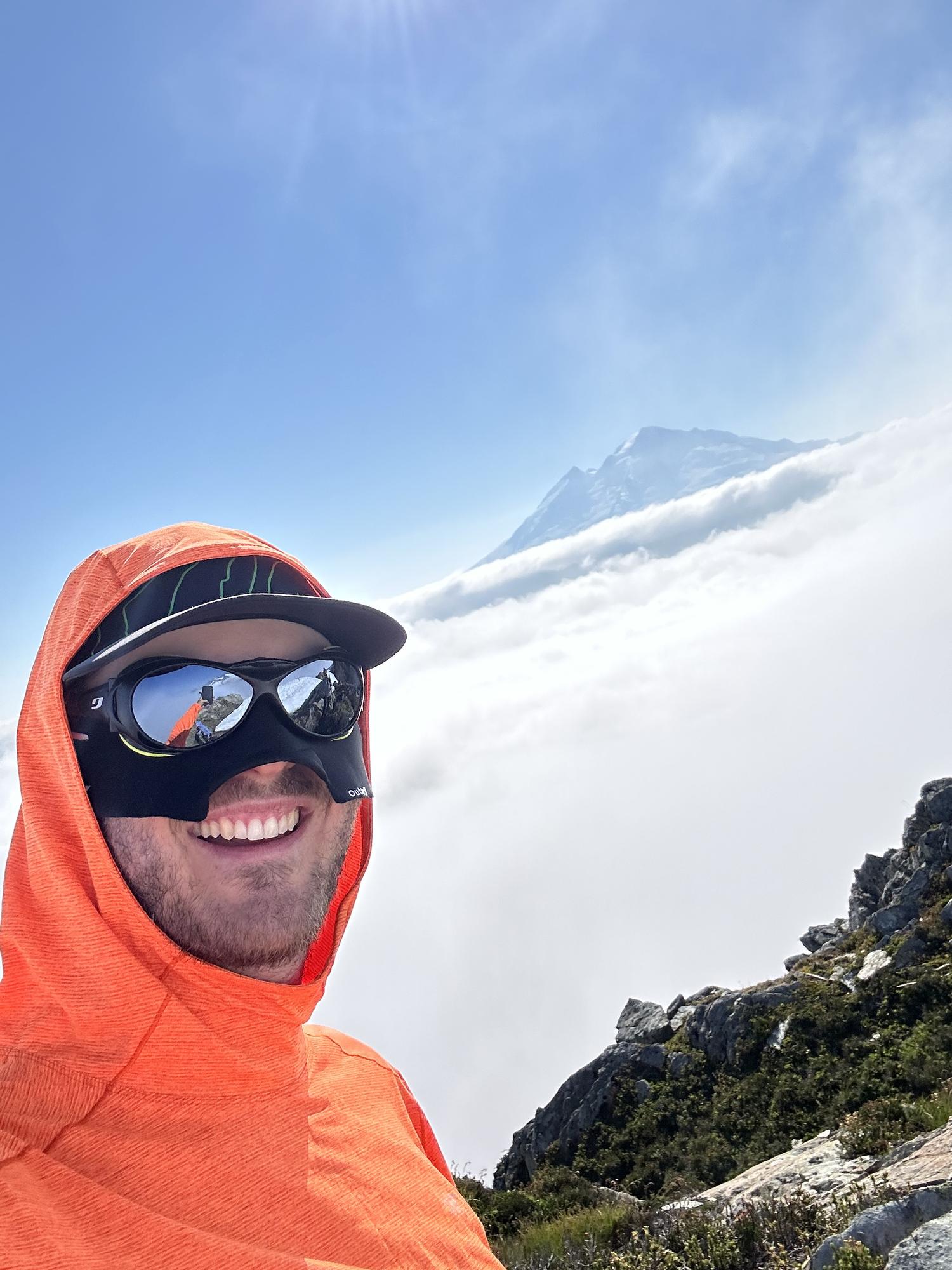 Jake Heller in an orange hooded sun shirt and glacier goggles above a thick cloud layer