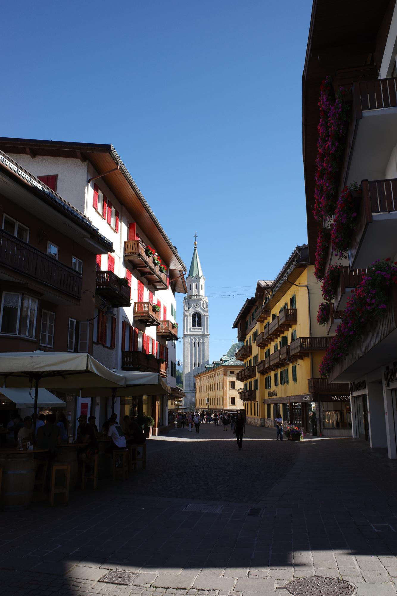 A sunlit pedestrian street in Cortina d'Ampezzo with a church bell tower at the end