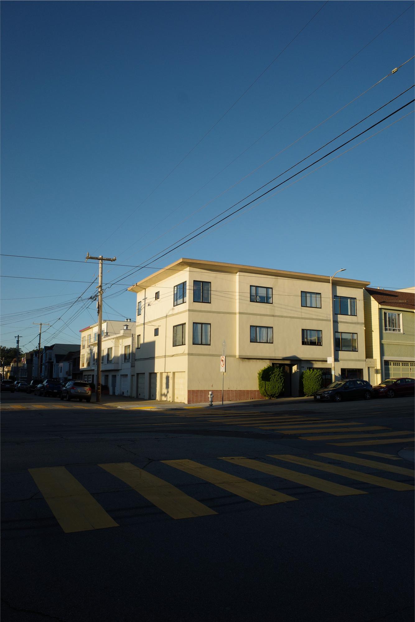 A cream-colored apartment building on a San Francisco corner lit by low golden-hour sun, crosswalk stripes in the foreground