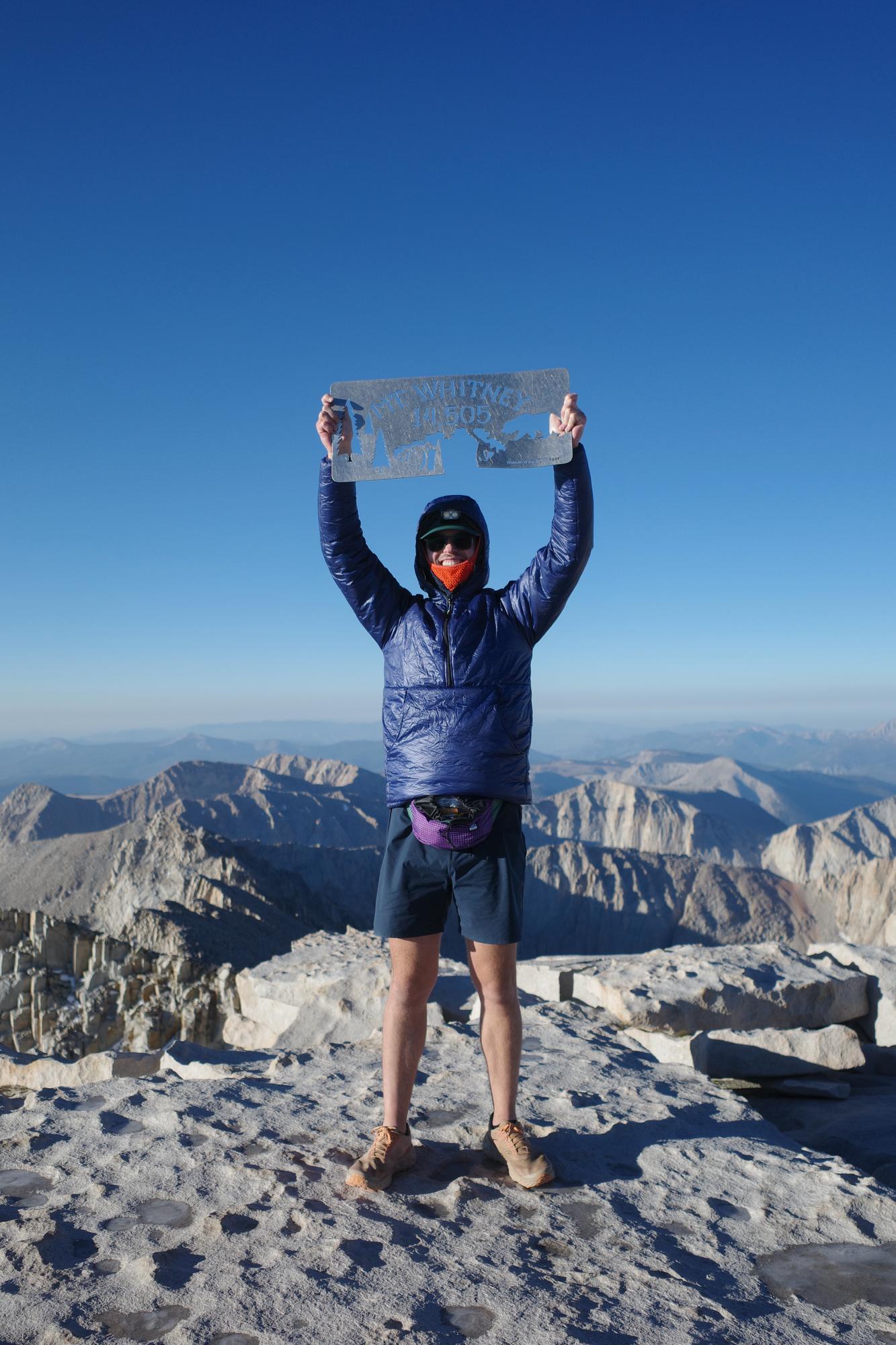 Jake Heller on a Sierra Nevada summit holding a sign overhead, mountains stretching to the horizon