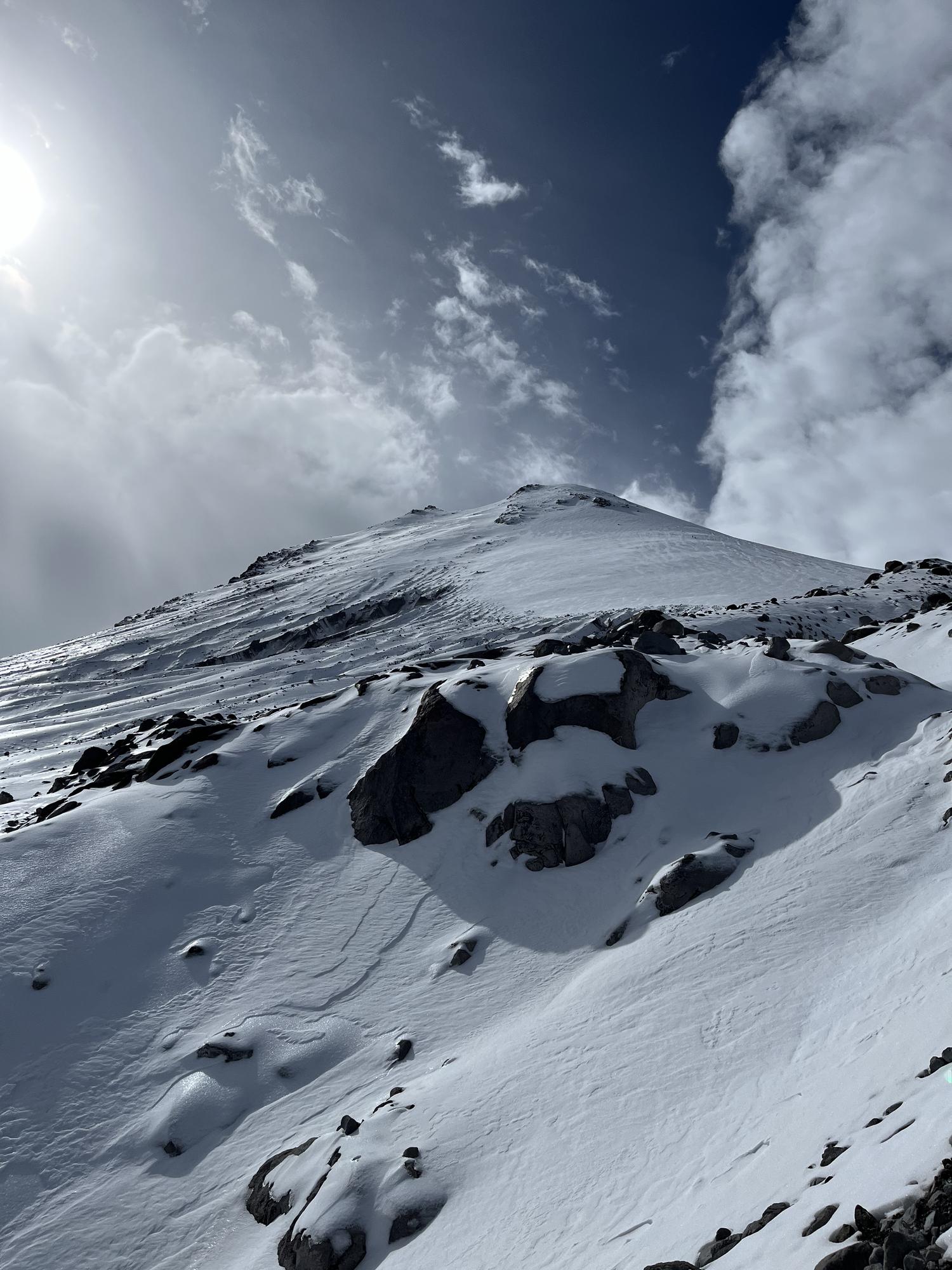 A snow slope climbing toward a corniced summit ridge with sun flare above
